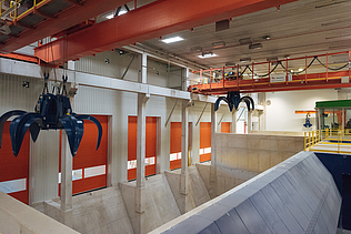 An indoor industrial facility featuring large blue overhead claw cranes positioned over concrete waste pits and orange loading bay doors.