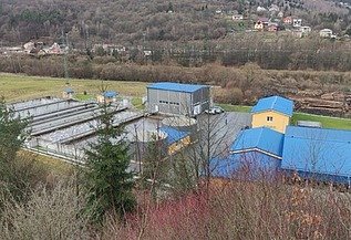 Aerial view of a water treatment facility with blue-roofed buildings and rectangular basins, surrounded by trees and hills in a rural area.