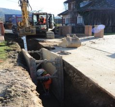 Excavator and workers installing a pipeline in a deep trench with protective shoring along a residential street.