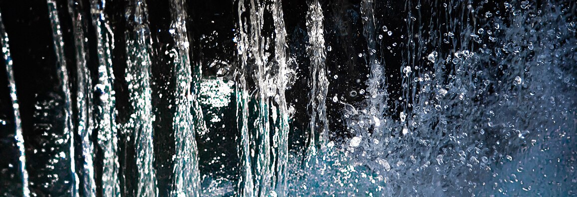 Nahaufnahme eines senkrecht fallenden Wasserstrahls mit spritzenden und im Licht schimmernden Tropfen vor einem dunklen Hintergrund.