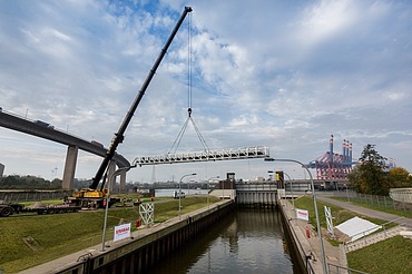 A mobile crane lifts a steel walkway over a lock at the Hamburg Harbor. The Köhlbrand Bridge and red container cranes are visible in the background, with STRABAG construction banners in the foreground.
