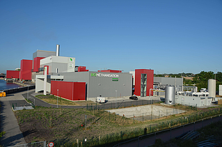 Diese Luftaufnahme zeigt die komplette Biomethanisierungsanlage mit ihren verschiedenen Gebäuden, Tanks und der Umgebung unter einem klaren blauen Himmel. An der rot-grauen Fassade ist der Schriftzug 