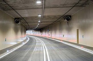 Curved, empty road tunnel with smooth concrete walls, ceiling lights, ventilation fans, and reflective lane markings.