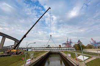 A large crane lifts a bridge section over a narrow canal near an industrial harbor.