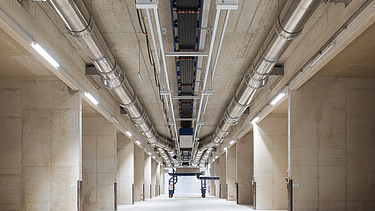 A long concrete corridor inside a facility with large silver metal pipes and a central conveyor system mounted to the ceiling.