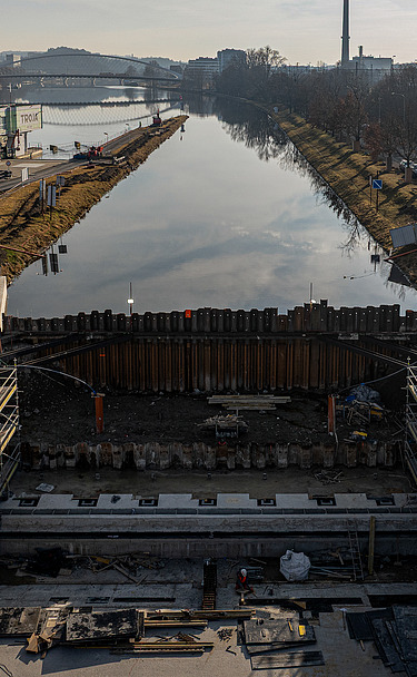 Construction site of a navigation channel lock along a river, showing reinforced concrete structures, steel sheet piling, and workers, with waterway extending into the background and urban buildings visible on the horizon.