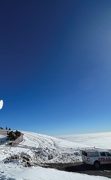 A telecommunications tower and a Strabag service vehicle on a snowy mountain summit above a sea of clouds.