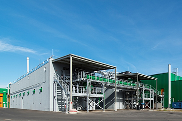 The image shows a modern industrial facility with green and gray buildings. In the foreground, there is an open steel structure with stairs, pipes, and technical equipment.