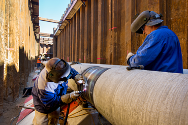 Two workers wearing protective gear are welding a large steel pipeline in a deep construction trench. The image shows precision pipeline installation, industrial engineering, and skilled labor in the energy infrastructure sector.