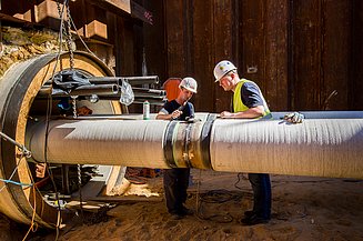 Two workers are inspecting and installing a large insulated pipe on an industrial construction site.
