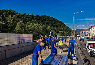 Group of workers in blue uniforms installing large metal panels along a roadside barrier. The scene includes a paved walkway, vehicles on the adjacent road, and green hills in the background under a clear blue sky.