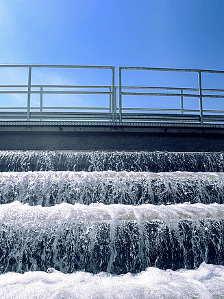 Mehrstufige Wasserkaskade in einer Kläranlage mit schäumendem Wasser, das über Stufen unter einer Metallgeländerkonstruktion unter blauem Himmel fließt.