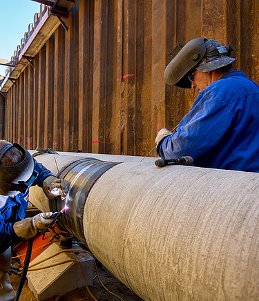 Two workers wearing protective gear are welding a large steel pipeline in a deep construction trench. The image shows precision pipeline installation, industrial engineering, and skilled labor in the energy infrastructure sector.