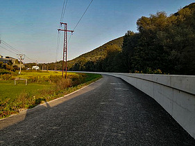 Straight paved road running alongside a tall concrete flood protection wall. On the left side, there is grassy terrain with power lines and poles, and a few houses in the distance. A forested hill rises on the right under a clear sky.