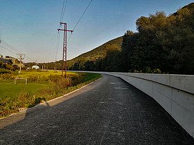 Gerade asphaltierte Straße, die an einer hohen Hochwasserschutzmauer aus Beton entlangführt. Auf der linken Seite befindet sich ein grasbewachsenes Gelände mit Stromleitungen und -masten sowie ein paar Häusern in der Ferne. Auf der rechten Seite erhebt sich ein bewaldeter Hügel unter klarem Himmel.
