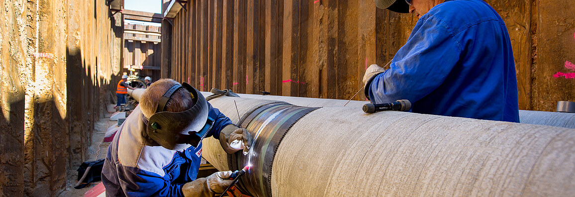 Two workers wearing protective gear are welding a large steel pipeline in a deep construction trench. The image shows precision pipeline installation, industrial engineering, and skilled labor in the energy infrastructure sector.