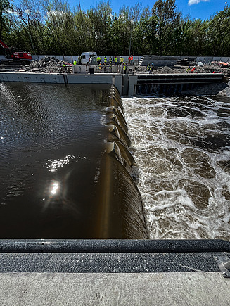 Nahaufnahme einer im Bau befindlichen Flussschleuse, die zeigt, wie das Wasser über eine Reihe von gebogenen Barrieren fließt, die eine kontrollierte Kaskade bilden, mit turbulentem Wasser auf der rechten Seite. Im Hintergrund sind Arbeiter in Schutzausrüstung und Baumaschinen entlang der Betonstruktur zu sehen, umgeben von Bäumen unter einem klaren blauen Himmel.