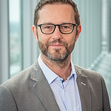 Person (Manfred) mit hellgrauer Anzugsjacke über einem hellblauen Hemd mit Kragen, steht in einem Innenraum vor hohen Glasfenstern, aus denen eine moderne Stadtlandschaft zu sehen ist.