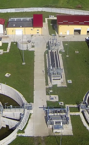 Luftaufnahme der Kläranlage von Moinești in Rumänien. Zu sehen sind runde Klärbecken, Behandlungsbecken, Kontrollgebäude mit roten Dächern und umliegende Grünflächen innerhalb eines eingezäunten Geländes.