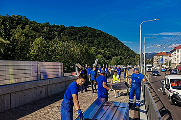 Group of workers in blue uniforms installing large metal panels along a roadside barrier. The scene includes a paved walkway, vehicles on the adjacent road, and green hills in the background under a clear blue sky.