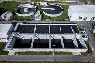 Aerial view of a modern wastewater treatment plant featuring rectangular sedimentation tanks in the foreground and two large circular clarifiers in the background, surrounded by green grass and facility buildings.