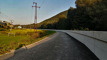 Straight paved road running alongside a tall concrete flood protection wall. On the left side, there is grassy terrain with power lines and poles, and a few houses in the distance. A forested hill rises on the right under a clear sky.