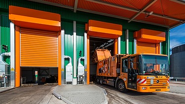 An orange refuse truck is shown at a modern recycling center with green and orange industrial bays. The vehicle is positioned at an open loading dock for waste disposal.