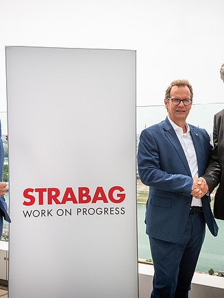 STRABAG and EVN/WTE executives shaking hands at the STRABAG headquarters in Vienna to mark the €100 million acquisition of WTE Wassertechnik GmbH. The men stand in front of a 