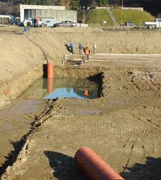 Construction site with workers installing large orange drainage pipes and a rectangular excavation filled with water.