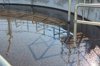 Nahaufnahme eines runden Wasseraufbereitungstanks, der teilweise mit dunklem Granulat und Wasser gefüllt ist. Entlang des Randes sind Edelstahlrohre und -geländer zu sehen, wobei sich die Struktur auf der Wasseroberfläche spiegelt.