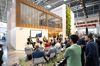 Visitors attending a panel discussion at the STRABAG and WTE exhibition stand at IFAT, featuring a modern booth design with wood elements, greenery, and a presentation area.