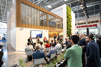 Besucher einer Podiumsdiskussion auf dem Messestand von STRABAG und WTE auf der IFAT, der sich durch ein modernes Standdesign mit Holzelementen, Begrünung und einem Präsentationsbereich auszeichnet.