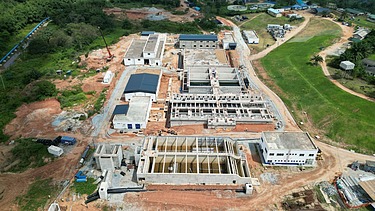 Aerial view of a large water treatment plant under construction, featuring multiple concrete structures, tanks, and surrounding green landscape.