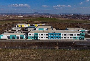 Luftaufnahme einer modernen Wasseraufbereitungsanlage in Prishtina mit mehreren niedrigen Gebäuden mit weißen und türkisfarbenen Fassaden, Flachdächern und umliegenden gepflasterten Straßen. Die Anlage befindet sich auf einer großen Freifläche, auf der im Hintergrund Baugeräte zu sehen sind, sowie in der Ferne Hügel unter einem teilweise bewölkten Himmel.