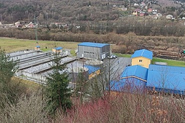 Aerial view of a water treatment facility with blue-roofed buildings and rectangular basins, surrounded by trees and hills in a rural area.