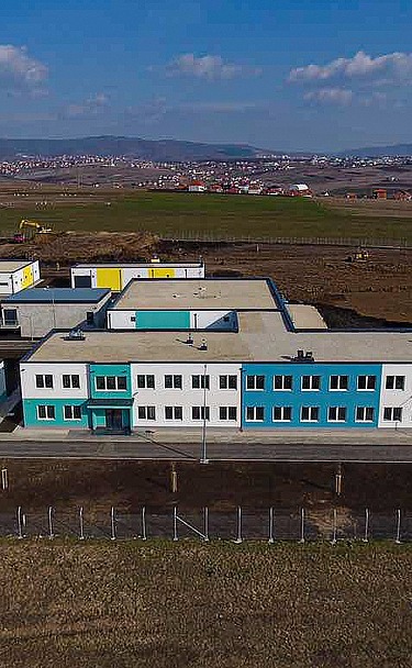 Aerial view of a modern water treatment plant in Prishtina, featuring multiple low-rise buildings with white and turquoise facades, flat roofs, and surrounding paved roads. The facility is located in a large open area with construction equipment visible in the background and distant hills under a partly cloudy sky.