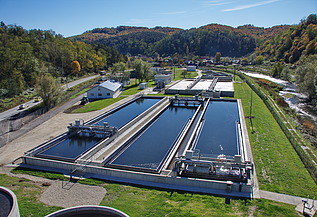 Aerial view of a wastewater treatment plant with multiple large rectangular basins, surrounded by green grass, trees, and rolling hills.
