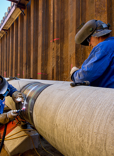 Two workers wearing protective gear are welding a large steel pipeline in a deep construction trench. The image shows precision pipeline installation, industrial engineering, and skilled labor in the energy infrastructure sector.