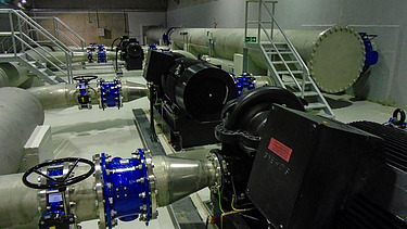 Interior view of an industrial facility with large pipelines, blue shut-off valves, and several black pump motors mounted on a concrete floor. Additional pipes and a metal staircase are visible in the background.
