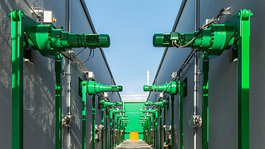 The image captures a symmetrical corridor between two industrial buildings, lined with green mechanical units mounted on sturdy supports.