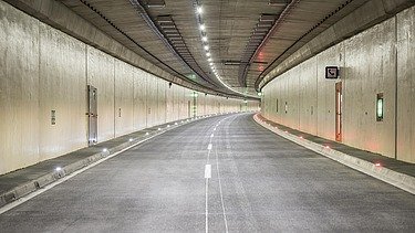 Eine asphaltierte Fahrbahn schlängelt sich in einem hell erleuchteten Betontunnel sanft in die Ferne. Oben beleuchten mehrere Reihen von LED-Leuchten den Weg, ergänzt durch weiße und rote Fahrbahnmarkierungsleuchten, die entlang der Gehwegkanten in den Belag integriert sind. Verschiedene Versorgungs- und Nottüren sind in die hellen Betonwände eingelassen.