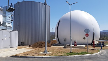 A close-up view of a wastewater treatment facility showing a large spherical biogas storage tank and a cylindrical anaerobic digester, with surrounding infrastructure and clear blue sky in the background.