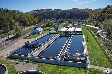 Aerial view of a wastewater treatment plant with multiple large rectangular basins, surrounded by green grass, trees, and rolling hills.