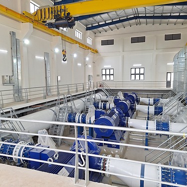 Interior view of a pumping station at the Xuan-Mai Surface Water Treatment Plant, featuring large blue pumps, interconnected pipelines, access platforms, and an overhead crane.