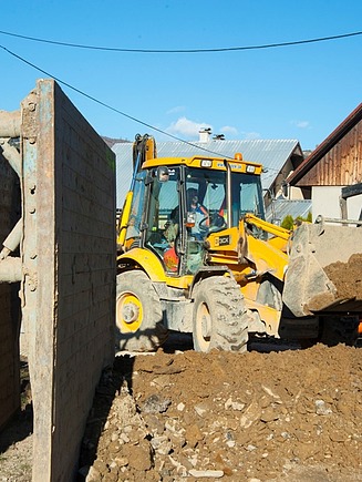 Yellow backhoe loader moving soil at a construction site with trench shoring panels and orange drainage pipes near residential houses.