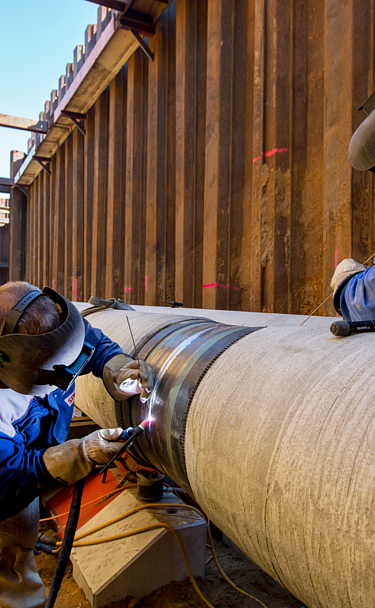 Two workers wearing protective gear are welding a large steel pipeline in a deep construction trench. The image shows precision pipeline installation, industrial engineering, and skilled labor in the energy infrastructure sector.