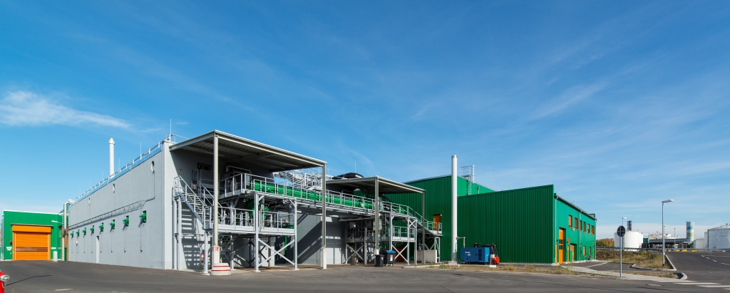 The image shows a modern industrial facility with green and gray buildings. In the foreground, there is an open steel structure with stairs, pipes, and technical equipment.
