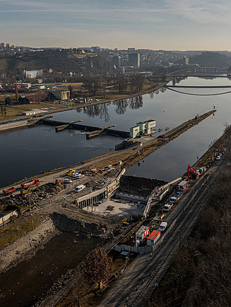 Luftaufnahme einer Flussschifffahrtskanal-Baustelle mit einem teilweise errichteten Schleusenbauwerk mit Stahlspundwänden, schweren Maschinen und Baufahrzeugen an den Ufern. Die Szene umfasst einen breiten Fluss mit Brücken im Hintergrund, angrenzende Bahngleise und eine hügelige Stadtlandschaft unter klarem Himmel.