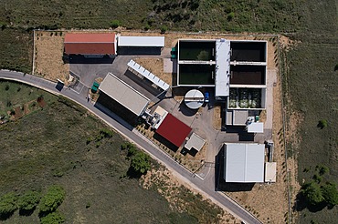 Top-down view of a wastewater treatment facility showing multiple rectangular treatment tanks, circular clarifier, and several buildings with red and white roofs, surrounded by green fields and access roads.