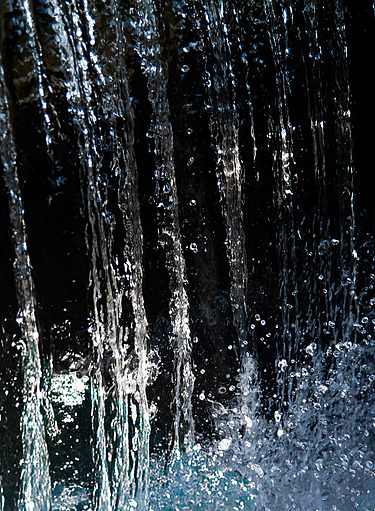 Nahaufnahme eines senkrecht fallenden Wasserstrahls mit spritzenden und im Licht schimmernden Tropfen vor einem dunklen Hintergrund.